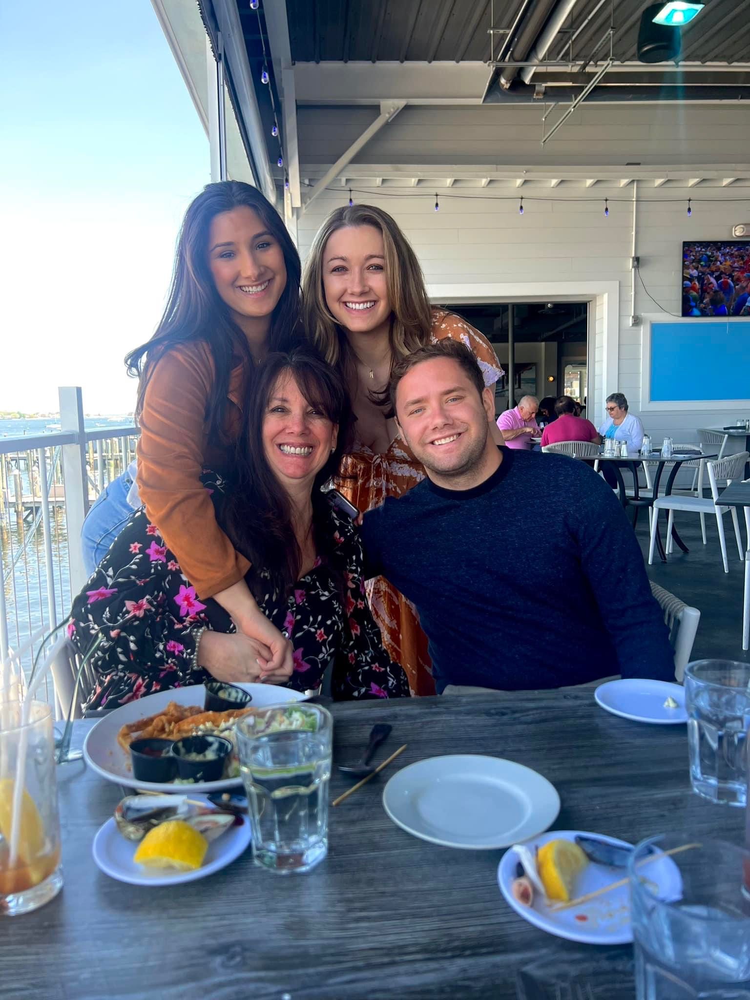 Four people smiling at outdoor restaurant table. Food and glasses visible.