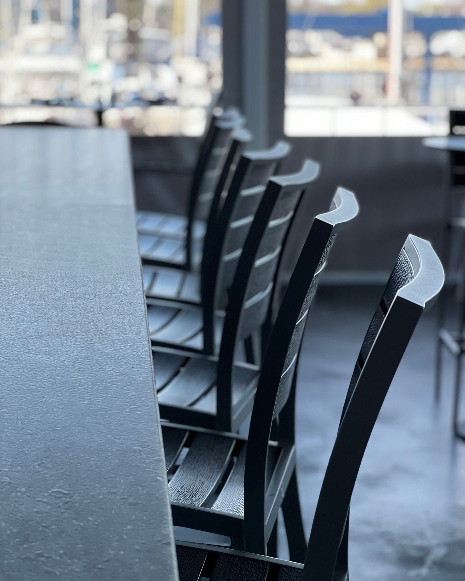 Row of black chairs lined up along a gray countertop in a waterfront setting.
