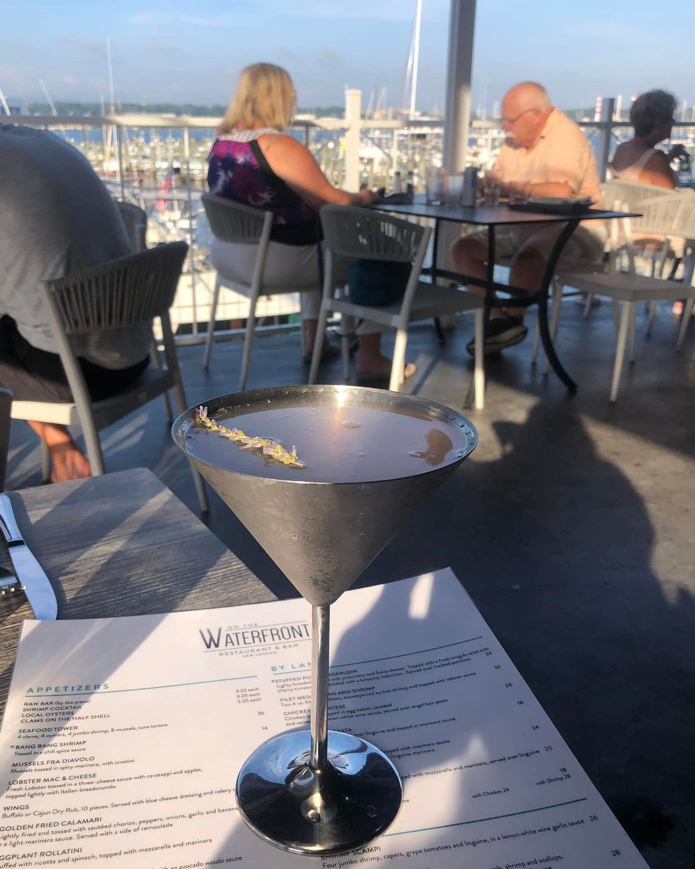 Cocktail in a metal martini glass on a table with a menu. People dine on an outdoor patio overlooking a marina.