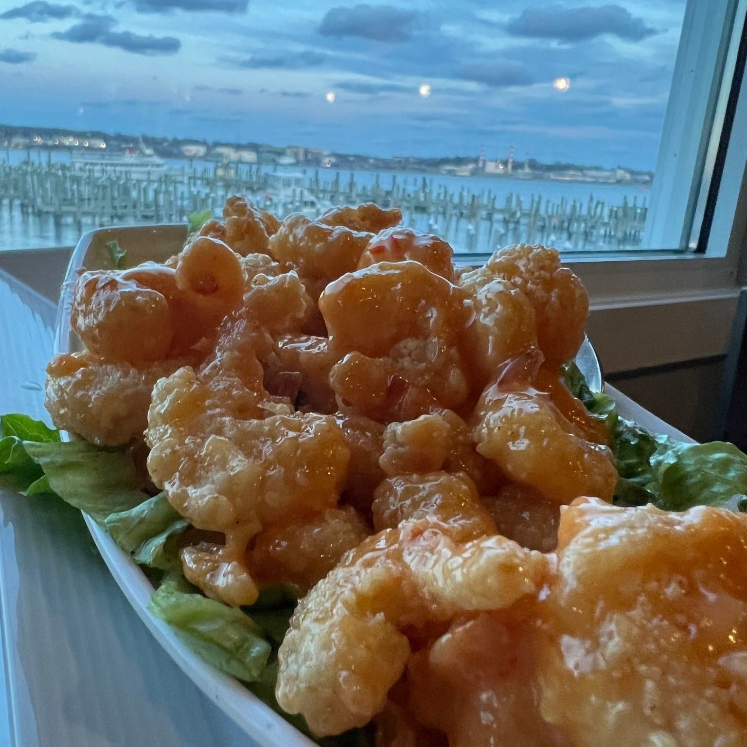 Plate of crispy fried shrimp with sauce on a bed of lettuce, with a harbor view in the background.
