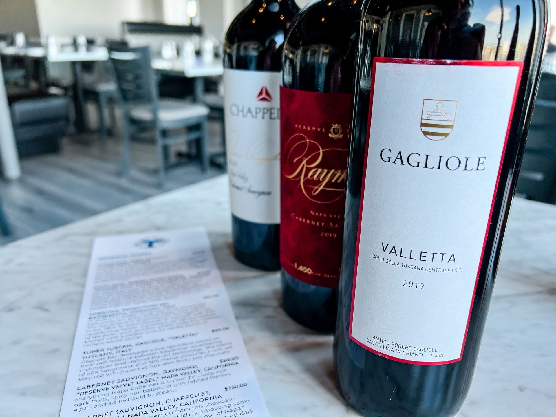 Three wine bottles on a table with a menu and restaurant background.