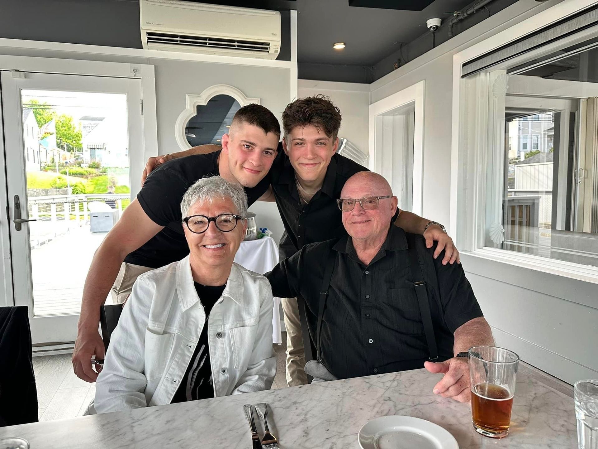 Family, two young men with their arms around older couple sitting at a table in a restaurant.