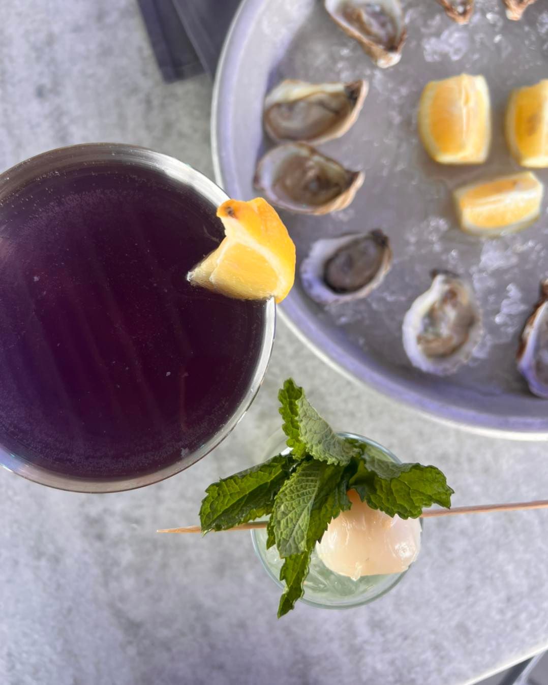 Cocktail with purple liquid, oyster plate with lemon wedges, mint garnish with lychee.