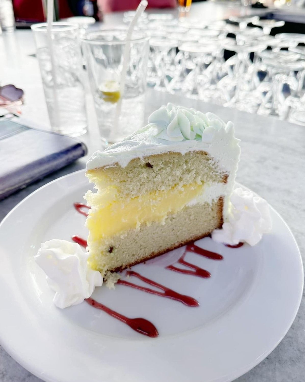 Slice of layered cake on a white plate with whipped cream and berry drizzle. Drinks in background.