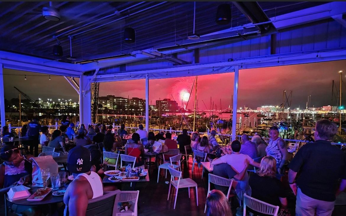 People watch fireworks from an outdoor restaurant at night. Red, white, and blue lights illuminate the crowd and sky.