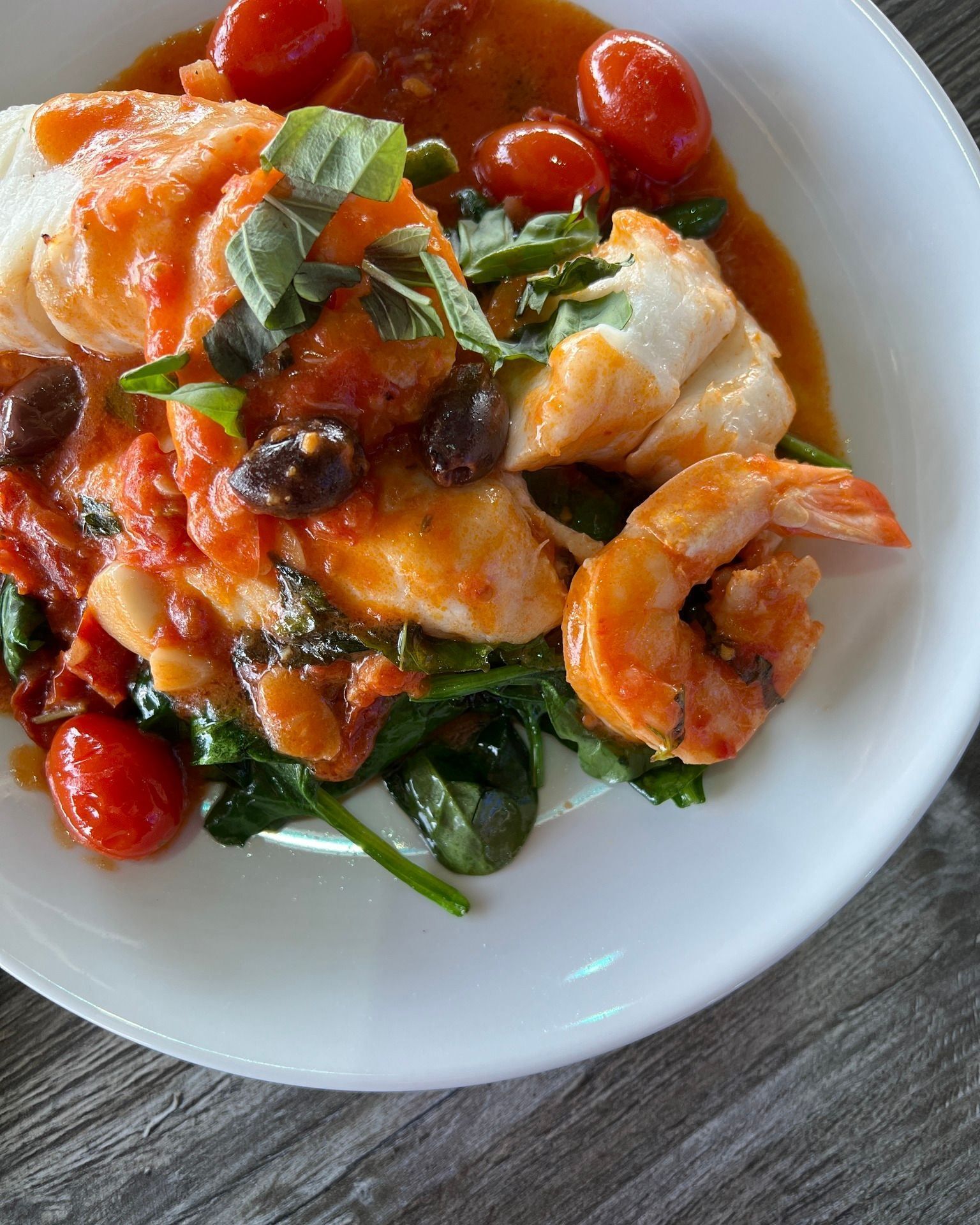 Plate of seafood: white fish, shrimp, tomatoes, olives, spinach, and sauce.