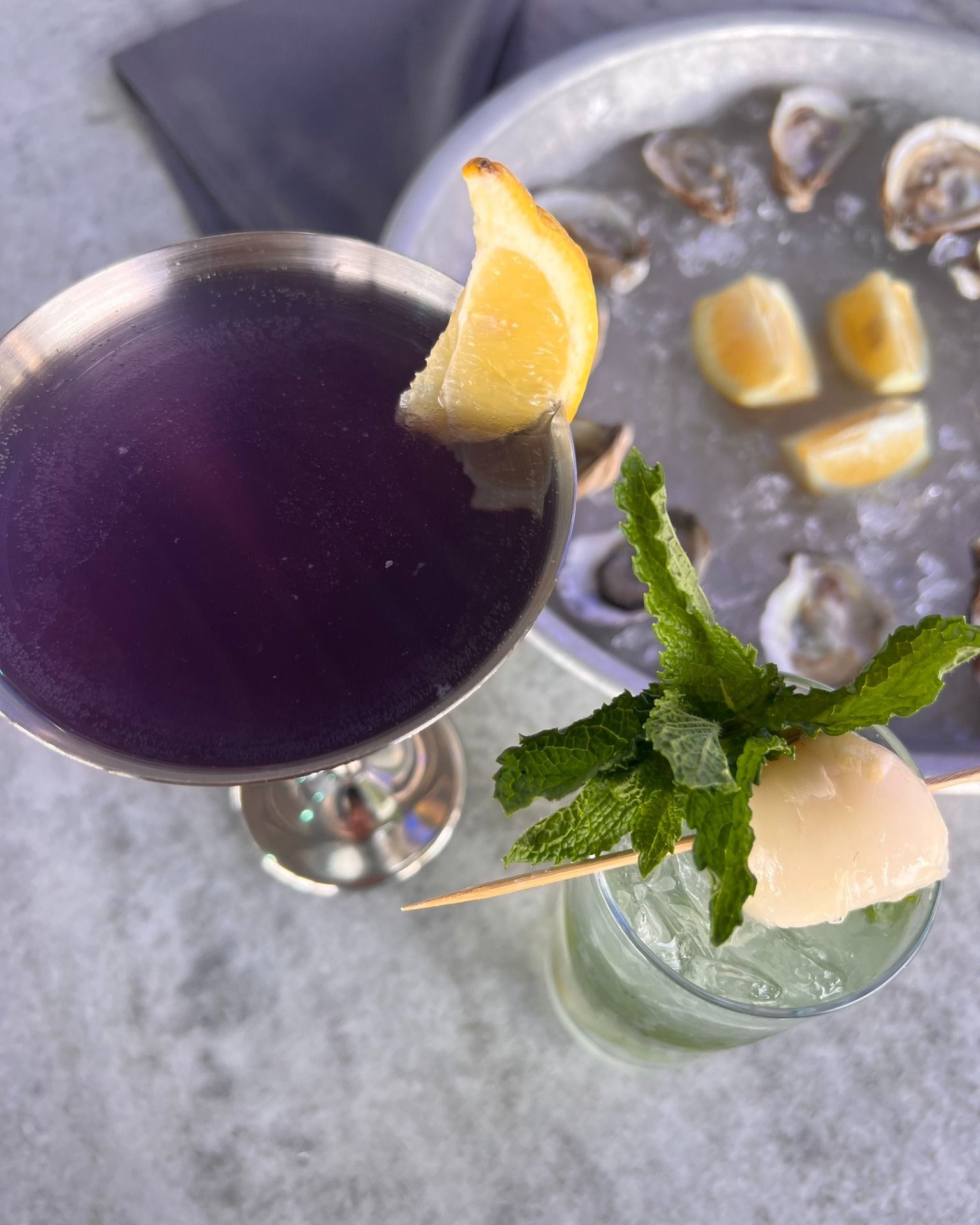 Purple cocktail with lemon garnish, green cocktail with lychee, oysters on ice.