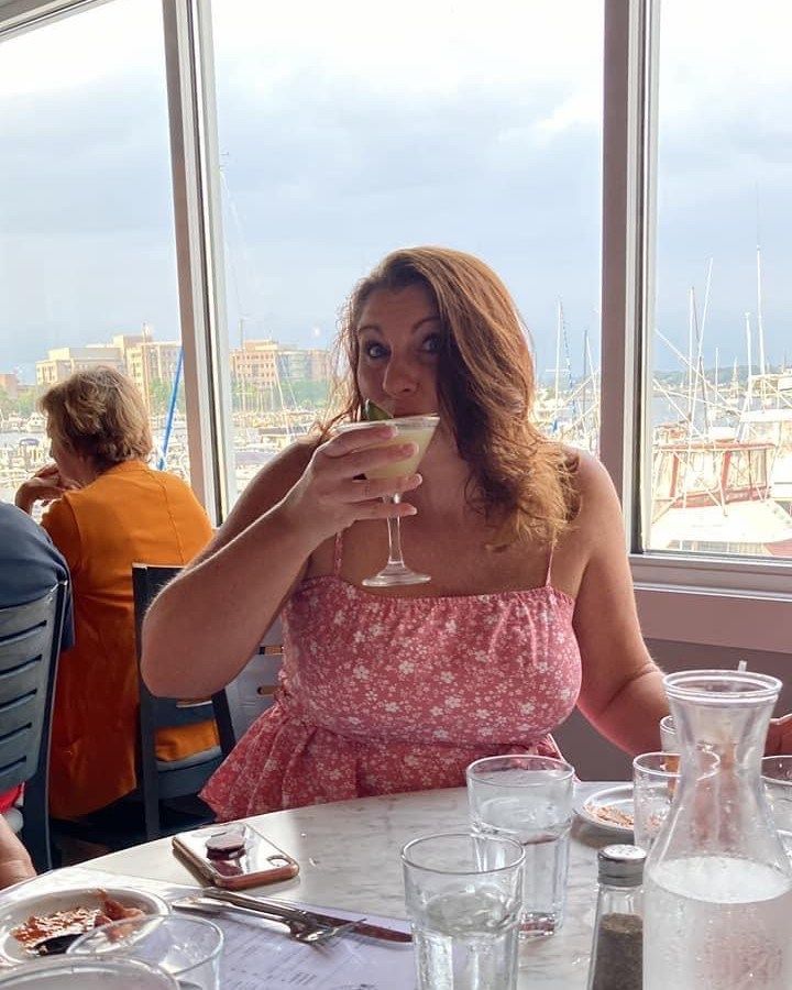 Woman sipping a cocktail at a restaurant overlooking a harbor. She wears a pink floral dress, has red hair, and smiles.