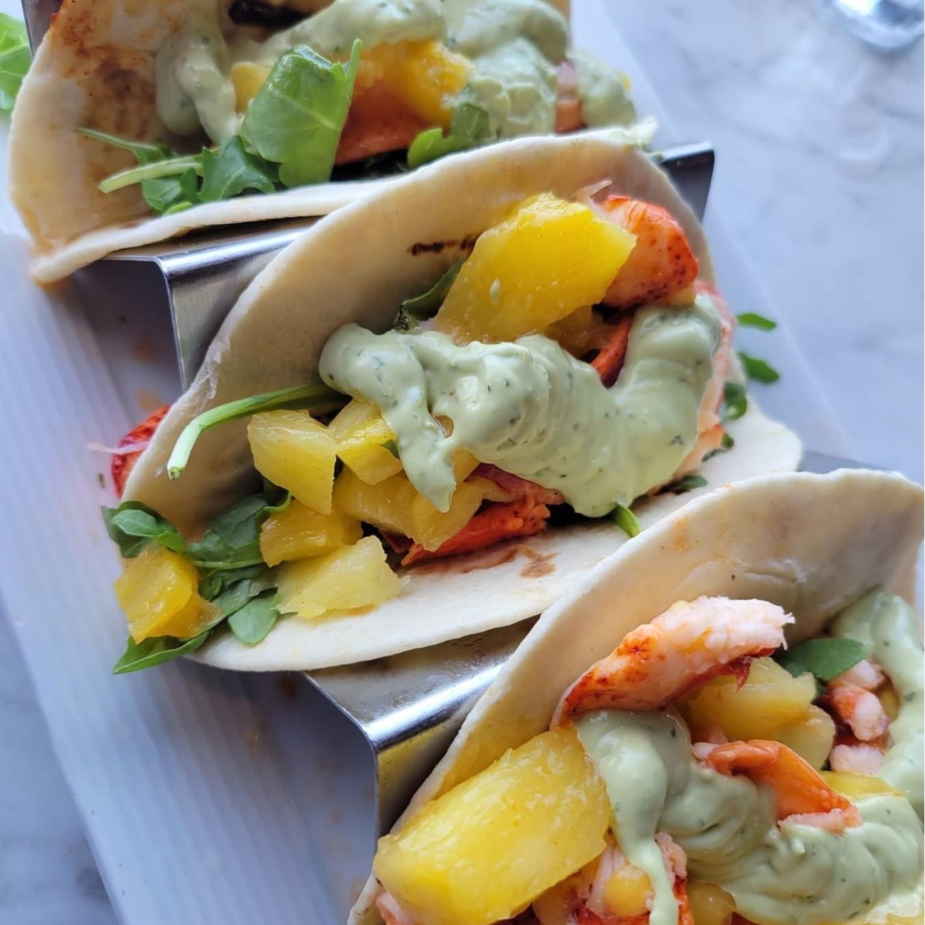 Three tacos with lobster, mango, arugula, and green sauce, served on a metal rack.