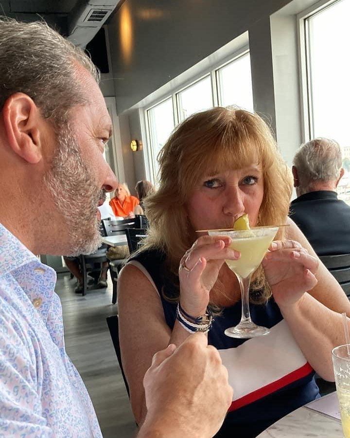 Woman sipping a cocktail with an olive garnish, seated near a man with a beard at a restaurant.