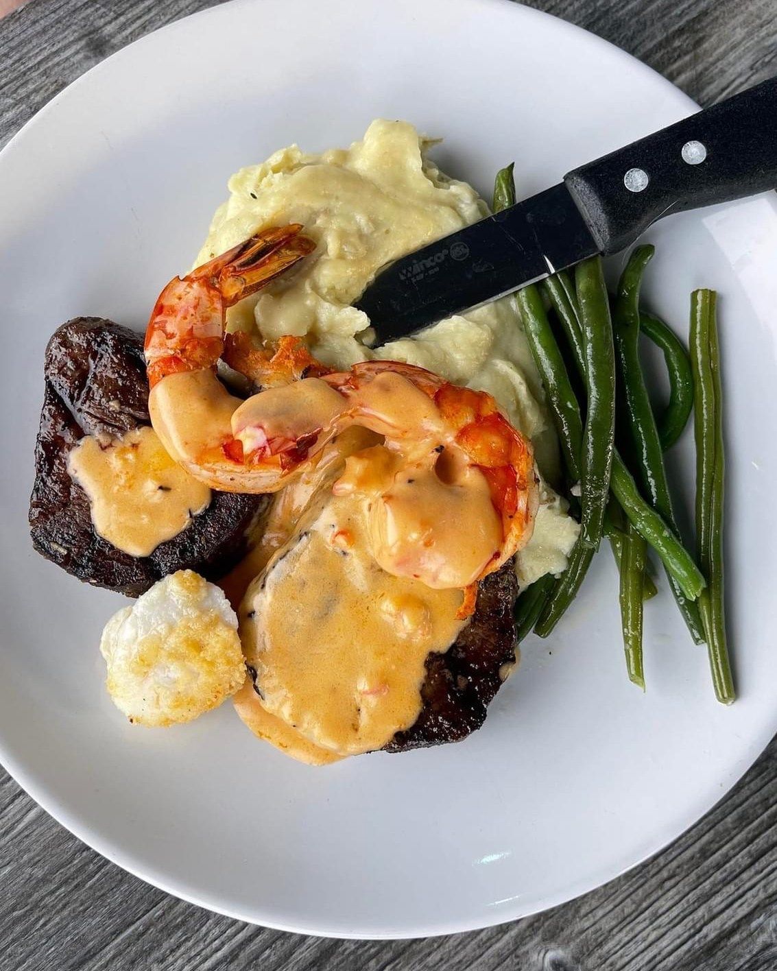 Steak, shrimp, mashed potatoes, and green beans on a white plate with a knife.