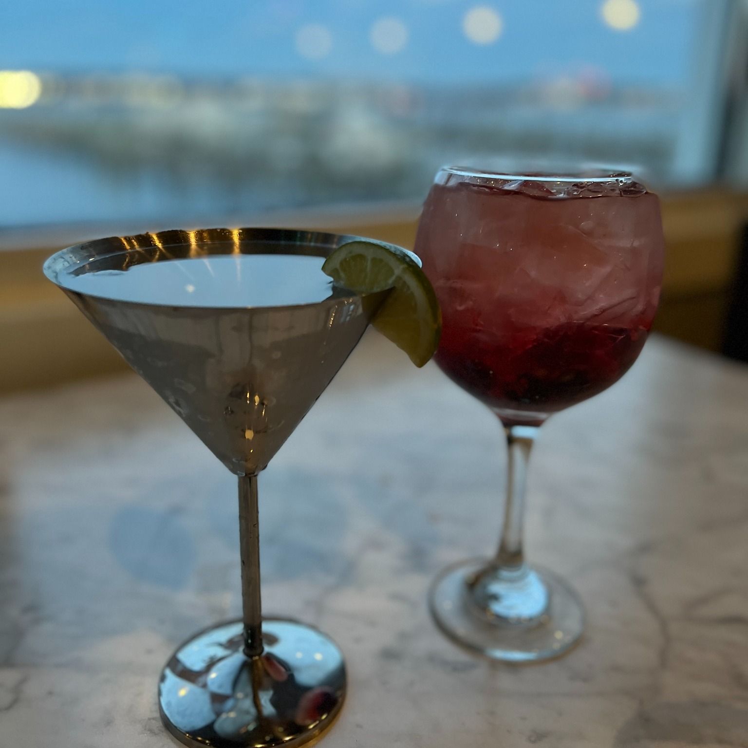 A silver martini with lime and a red berry drink in a wine glass, set before a window.