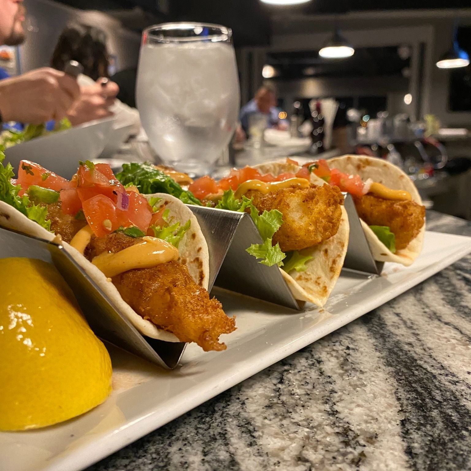 Fish tacos on a white plate with lemon, glass of water, and people in background at a restaurant.
