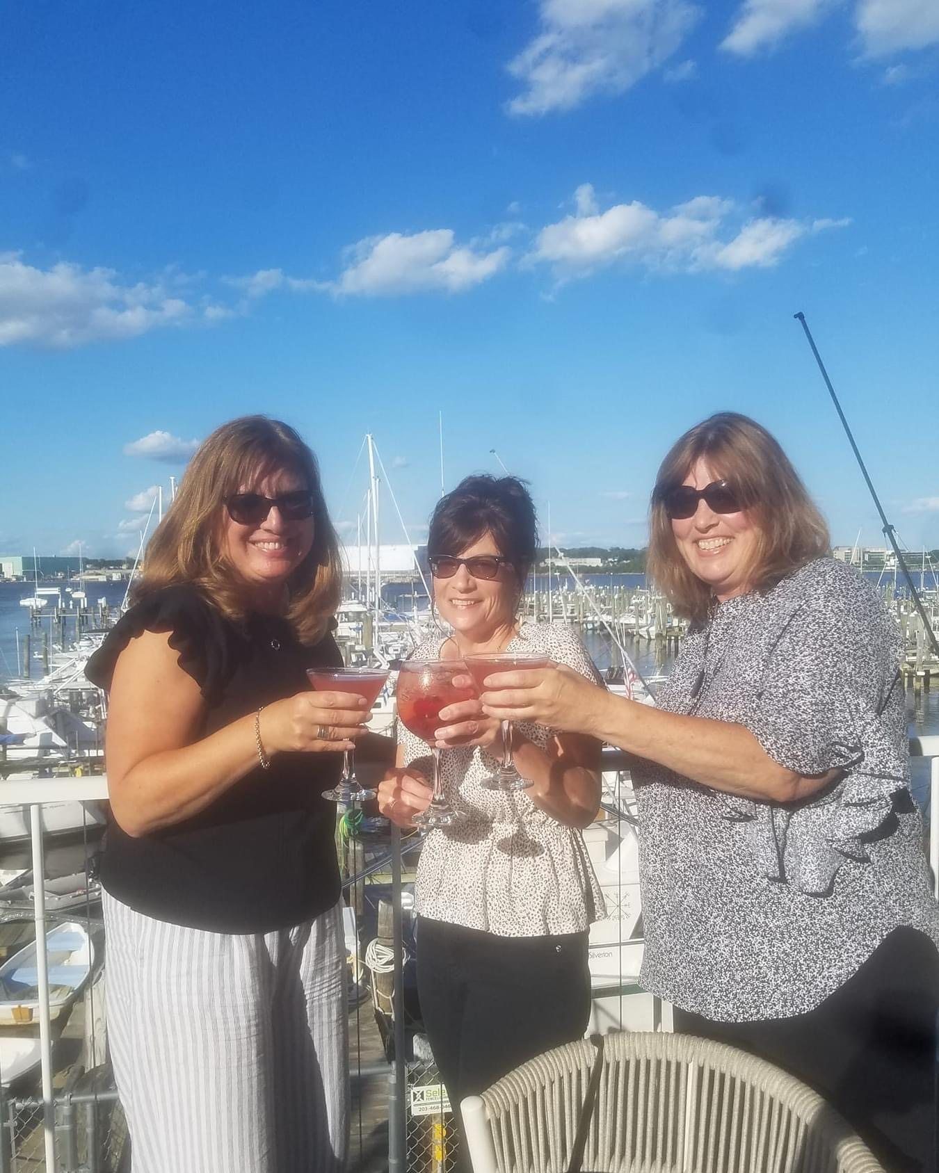 Three women toasting cocktails outdoors with a marina in the background under a blue sky.