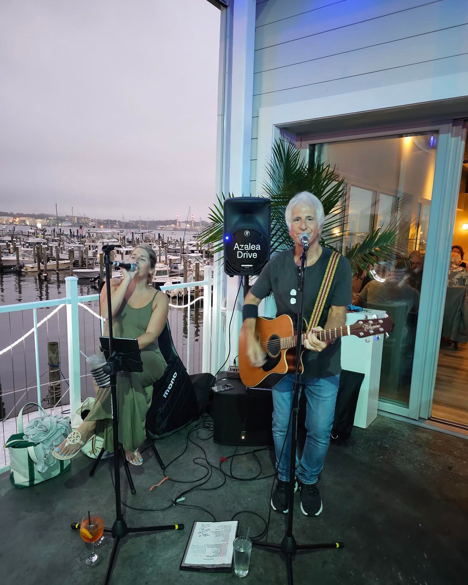 Musician plays acoustic guitar on outdoor patio, female singer next to him, water view.