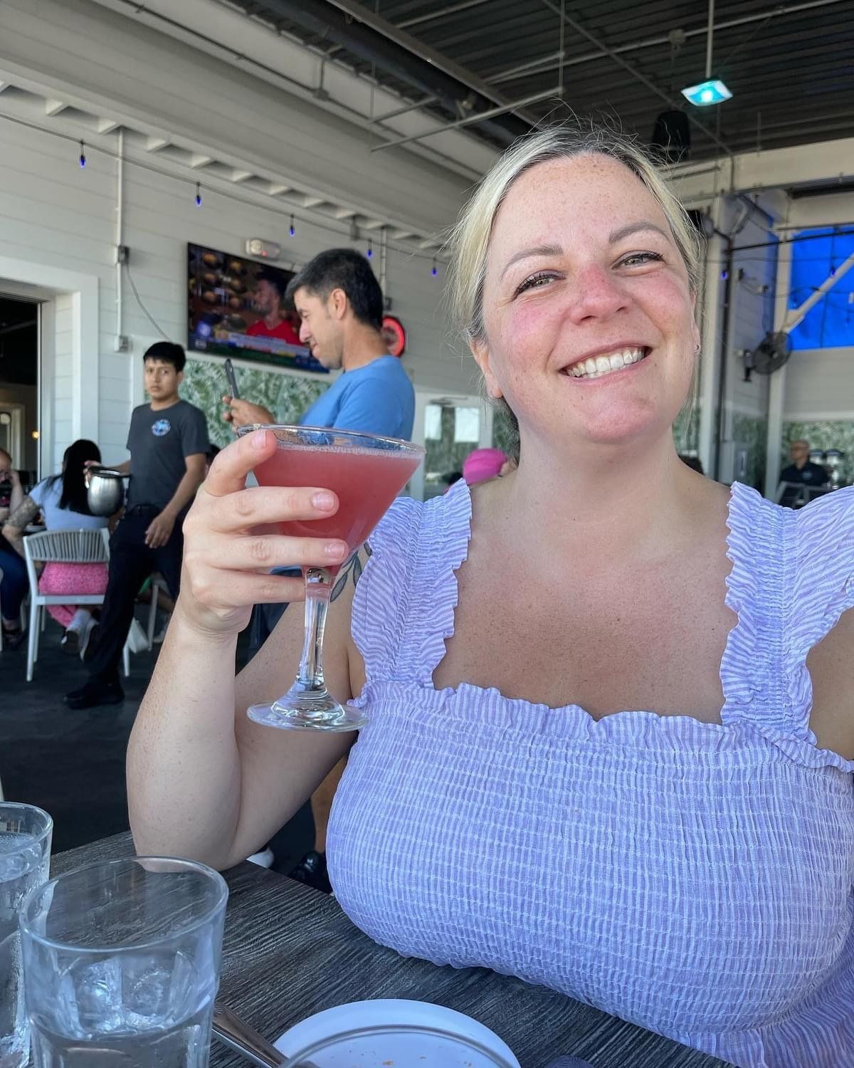 Woman smiling, holding a pink cocktail at an outdoor restaurant.