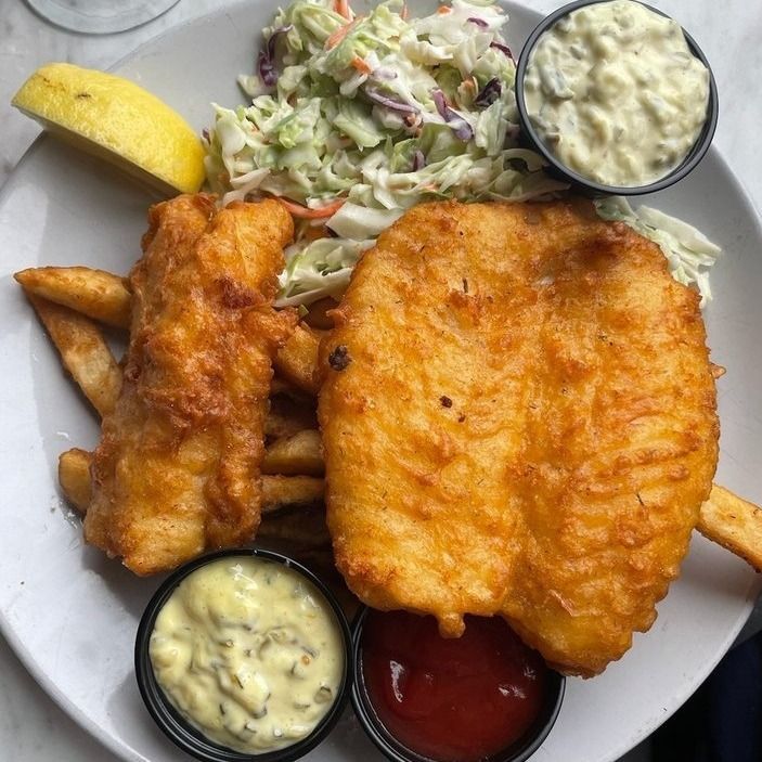 Plate of fish and chips: fried fish, fries, coleslaw, lemon wedge, and dipping sauces.