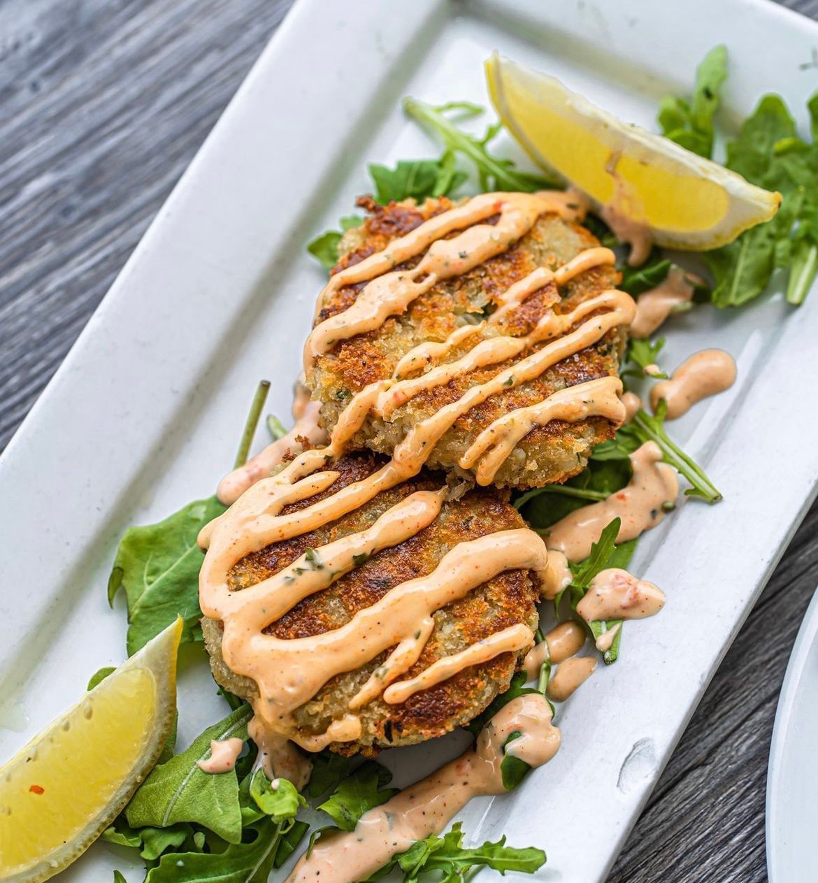 Two crab cakes drizzled with sauce on a bed of greens, garnished with lemon wedges.
