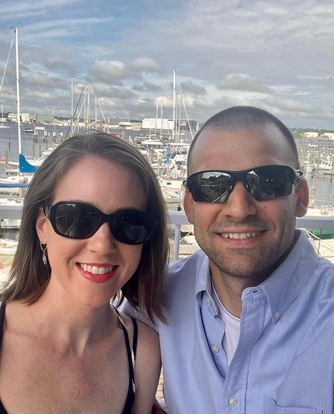 Couple smiling, wearing sunglasses, in front of a harbor with sailboats. Cloudy sky.