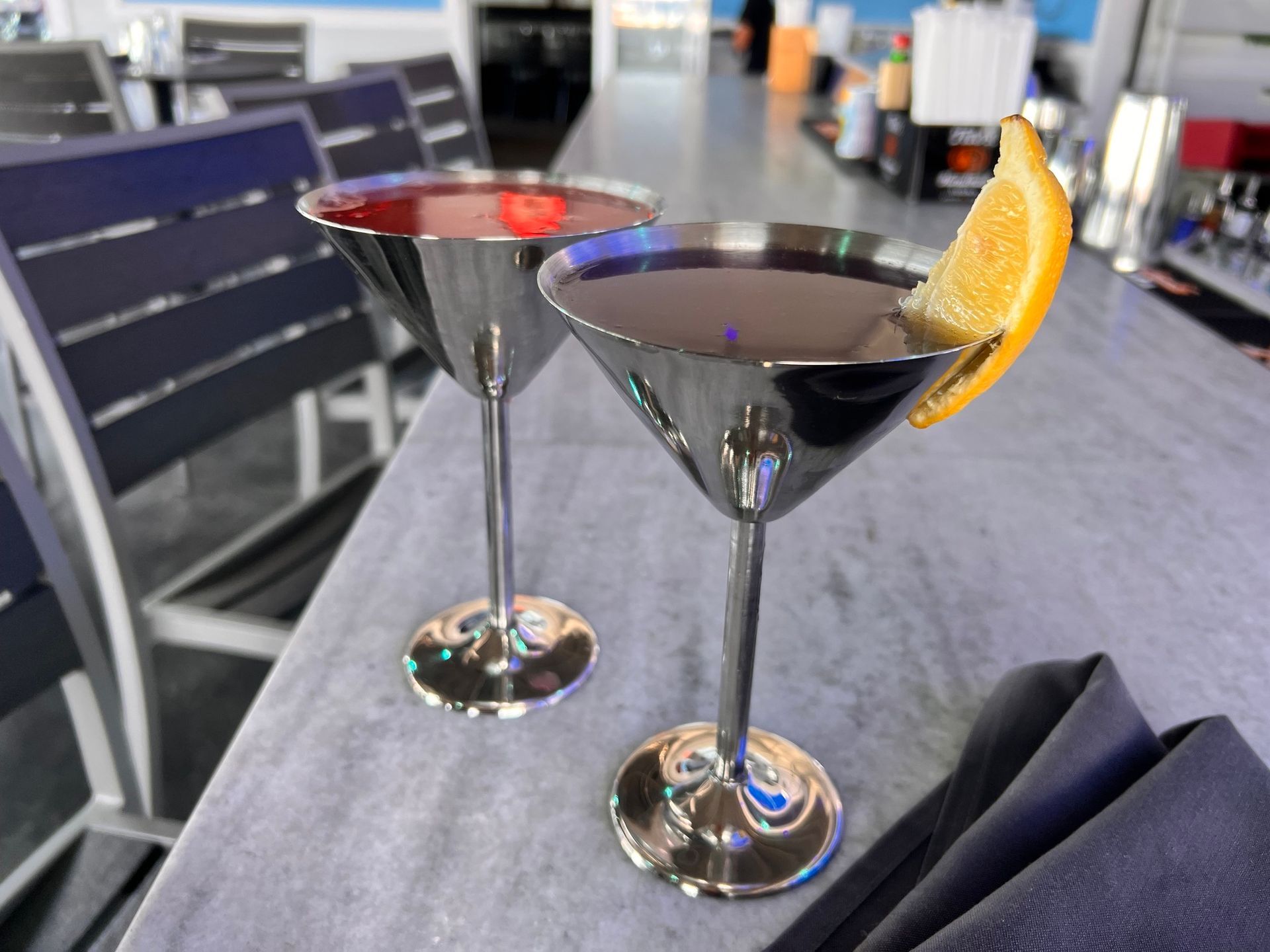 Two cocktails in silver martini glasses, one with an orange slice, on a bar.