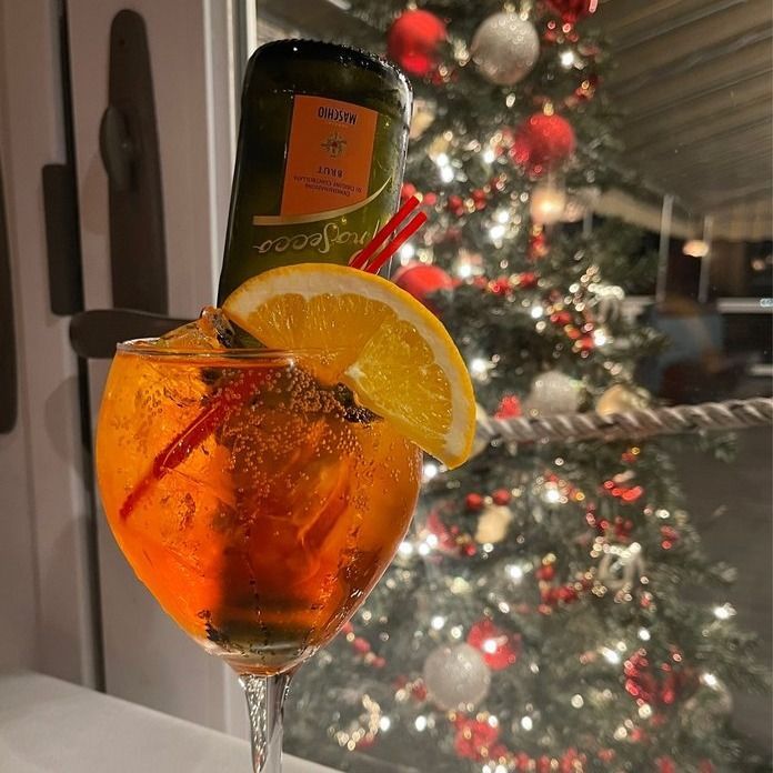 Aperol spritz cocktail with orange slice and straw, leaning against a prosecco bottle, holiday tree background.