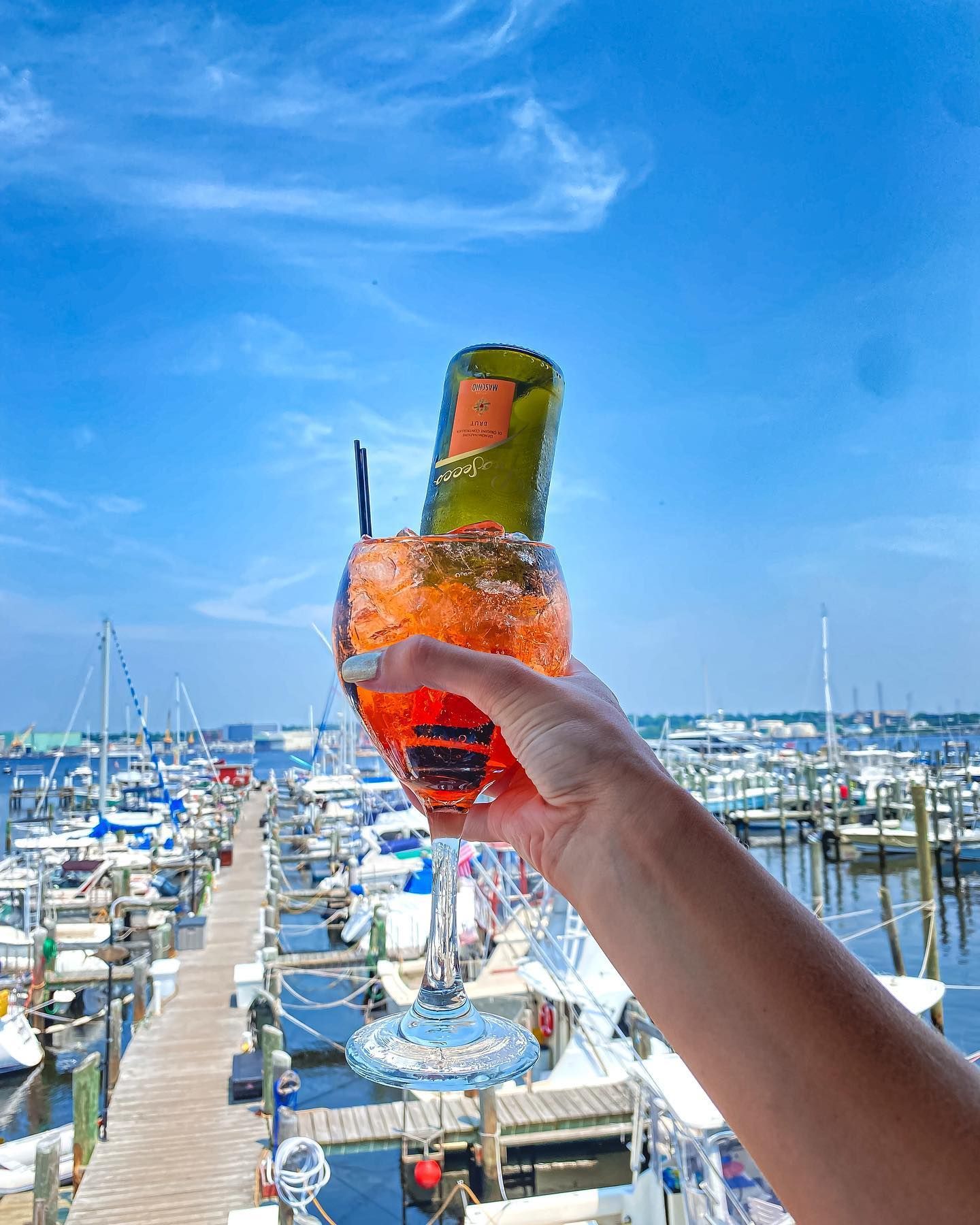 Hand holding a large cocktail glass with a small bottle, over a marina.