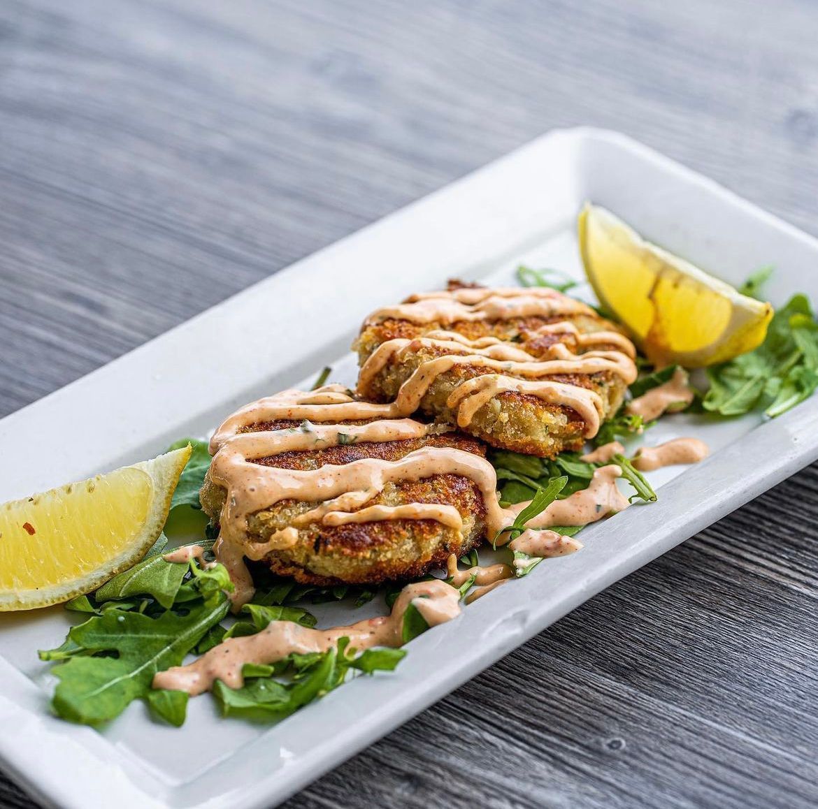 Two crab cakes drizzled with sauce on a bed of greens, served with lemon wedges on a white plate.