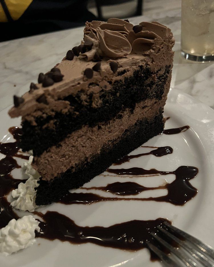 Slice of chocolate cake with chocolate frosting, chocolate syrup, and whipped cream on a white plate.