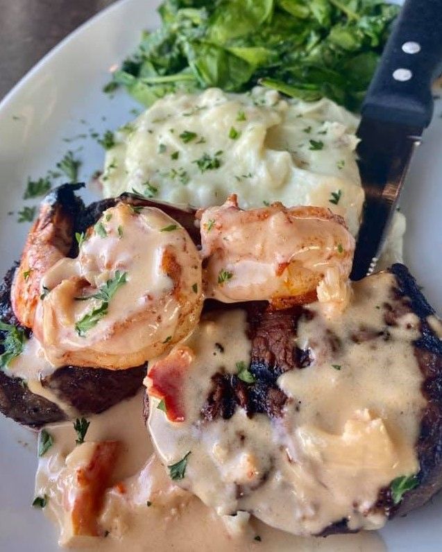 Steak and shrimp with creamy sauce, mashed potatoes, and spinach on a white plate.