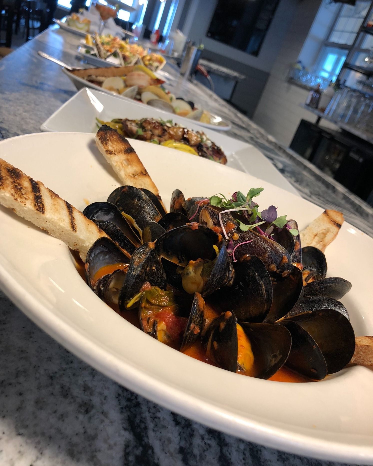 Mussels in a white bowl with grilled bread. Served on a marble countertop, other dishes in the background.