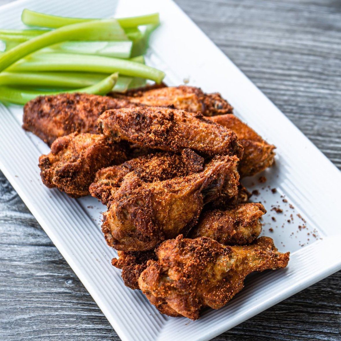 Plate of crispy, seasoned chicken wings with celery sticks on a gray wooden surface.