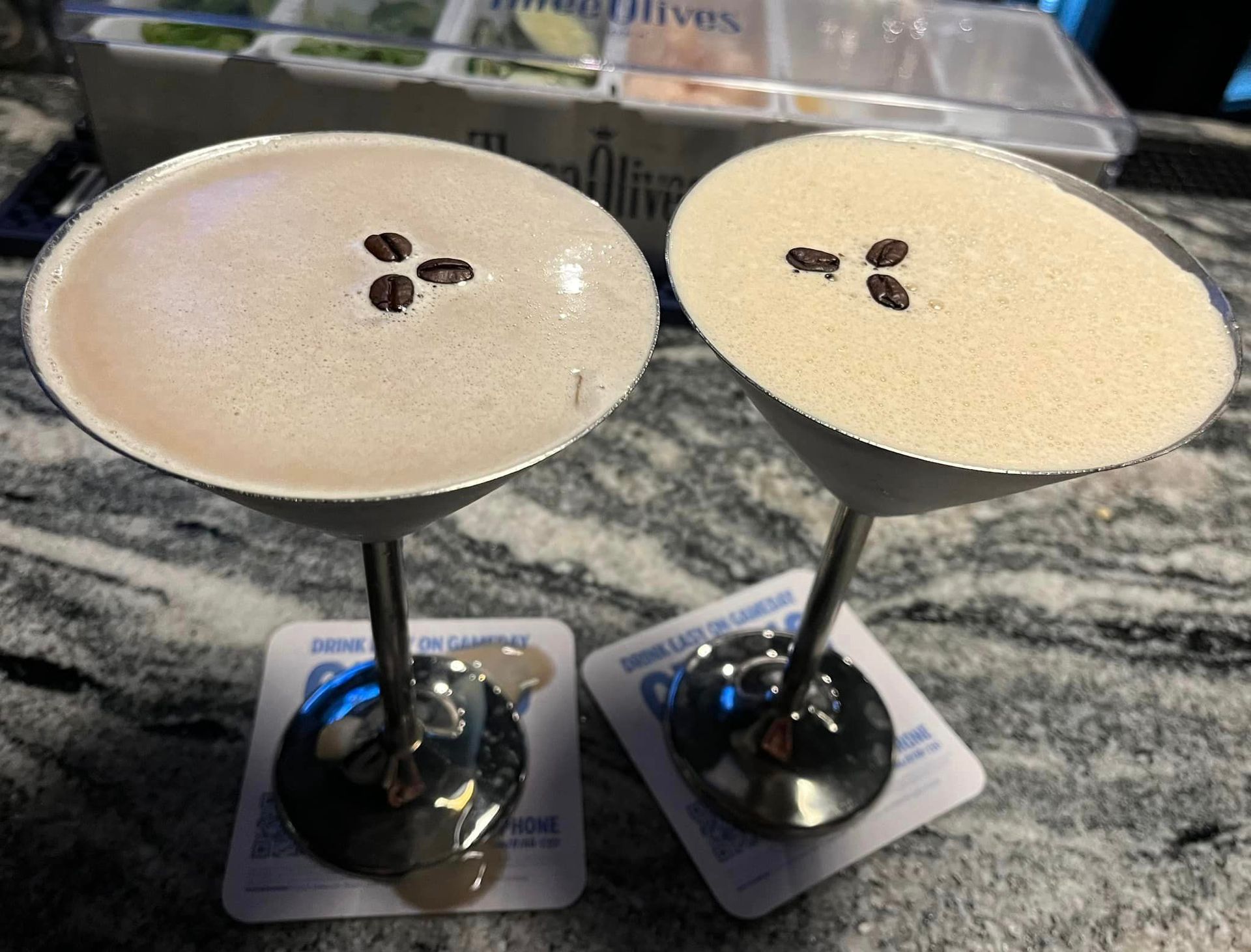Two coffee martinis with three coffee beans on a foamy top, on a bar with coasters.