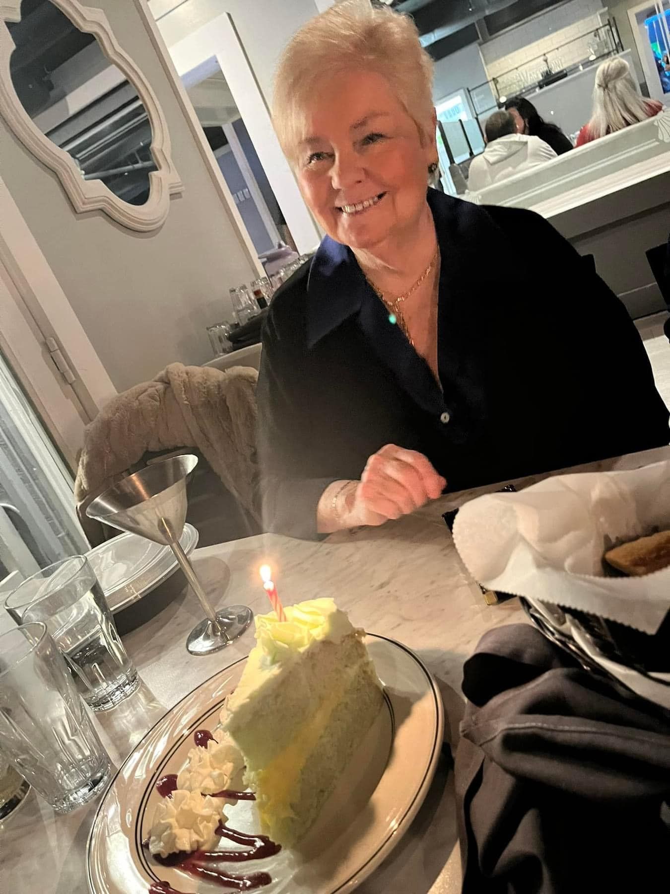 Woman smiles at a table with cake and a lit candle. Restaurant setting.