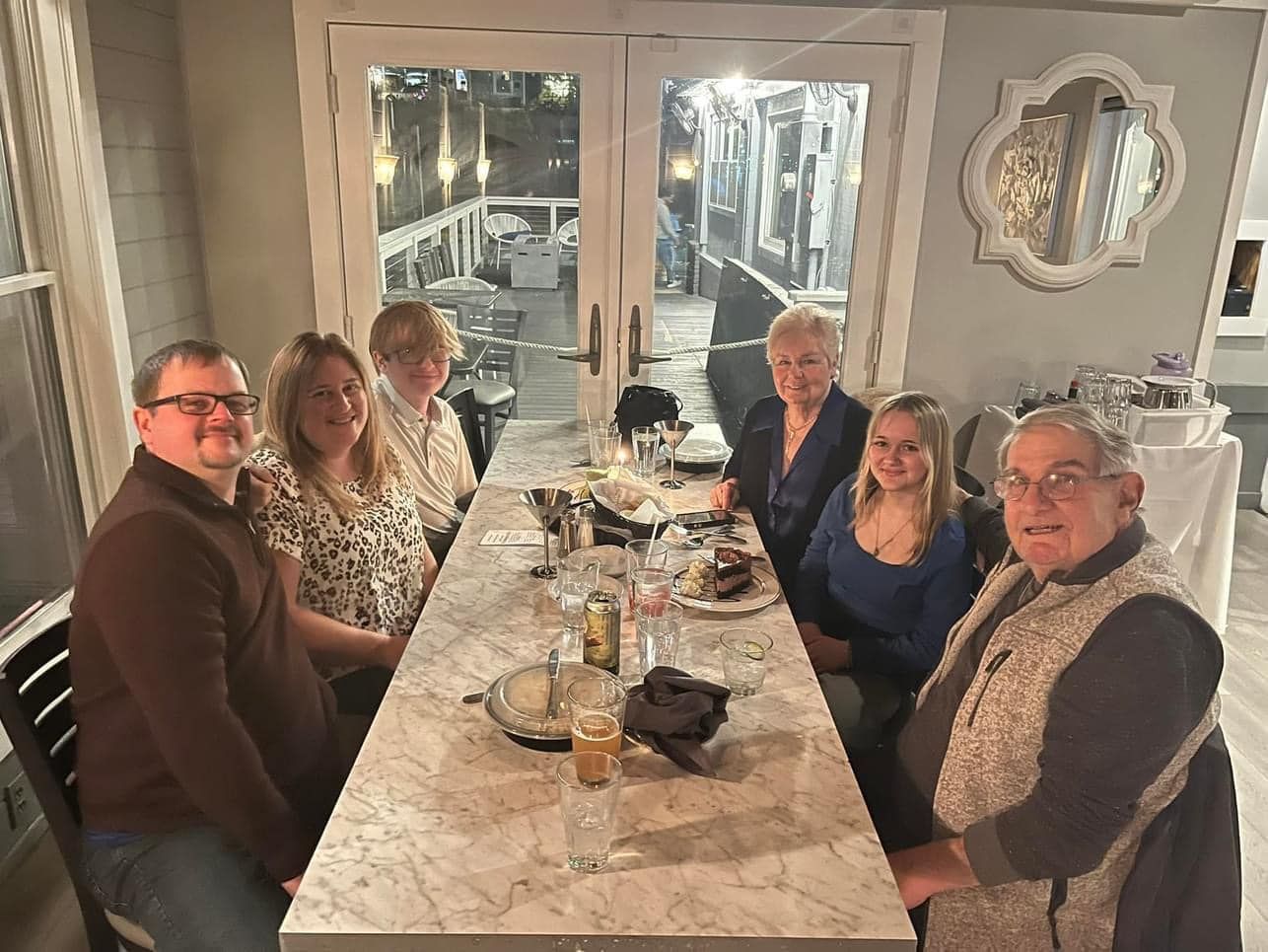 People seated at a table in a restaurant. Windows and a deck are visible in the background.