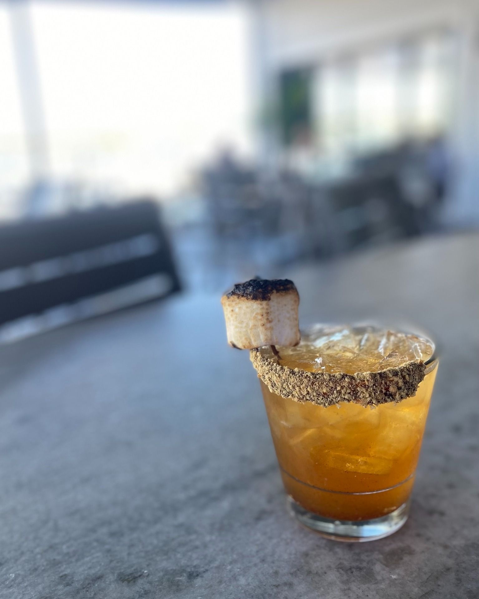 Cocktail in a glass with a dark rim and cake garnish, on a concrete surface outside.