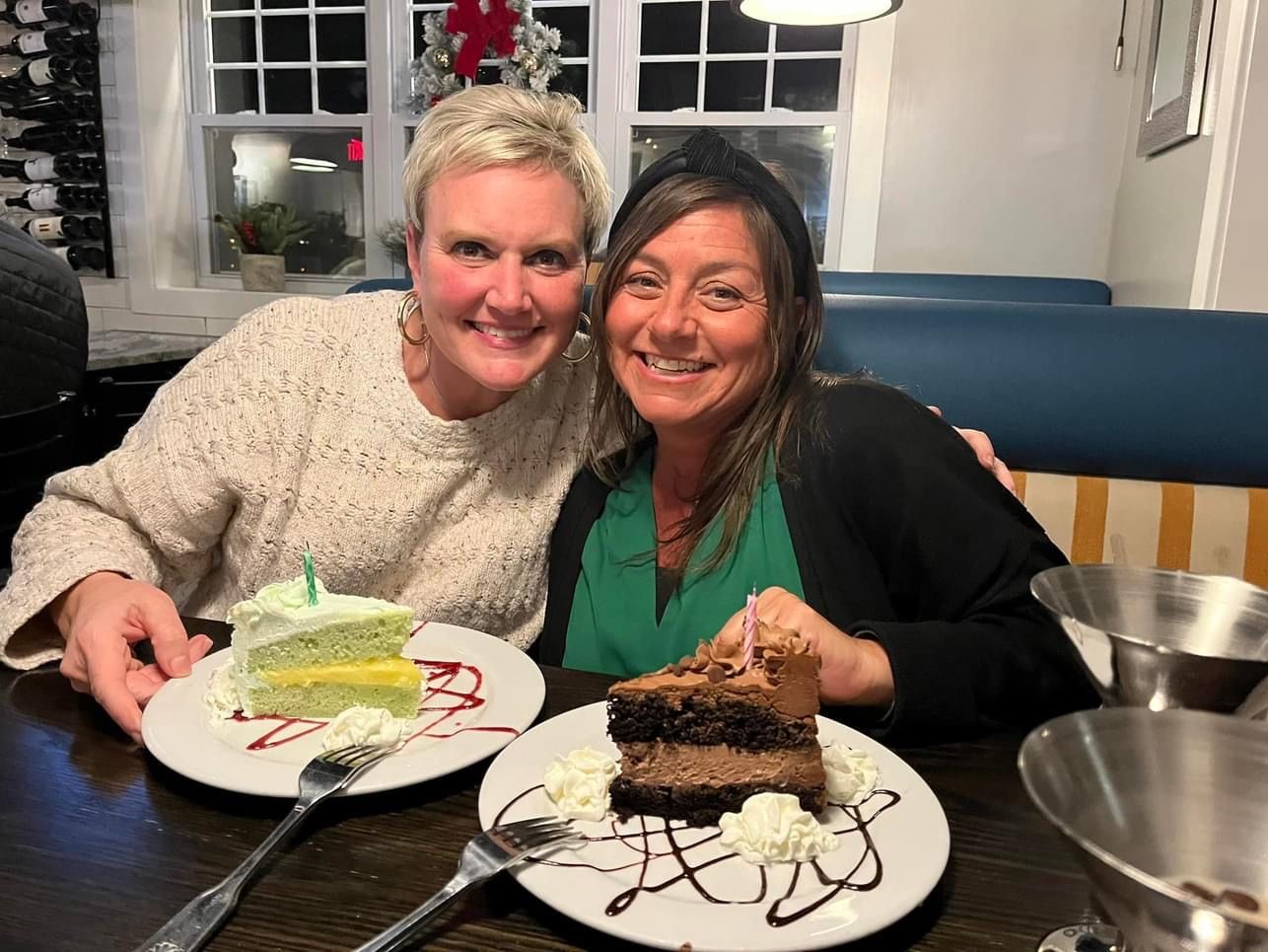 Two women smiling, holding plates of cake in a restaurant. One cake is green, the other chocolate.