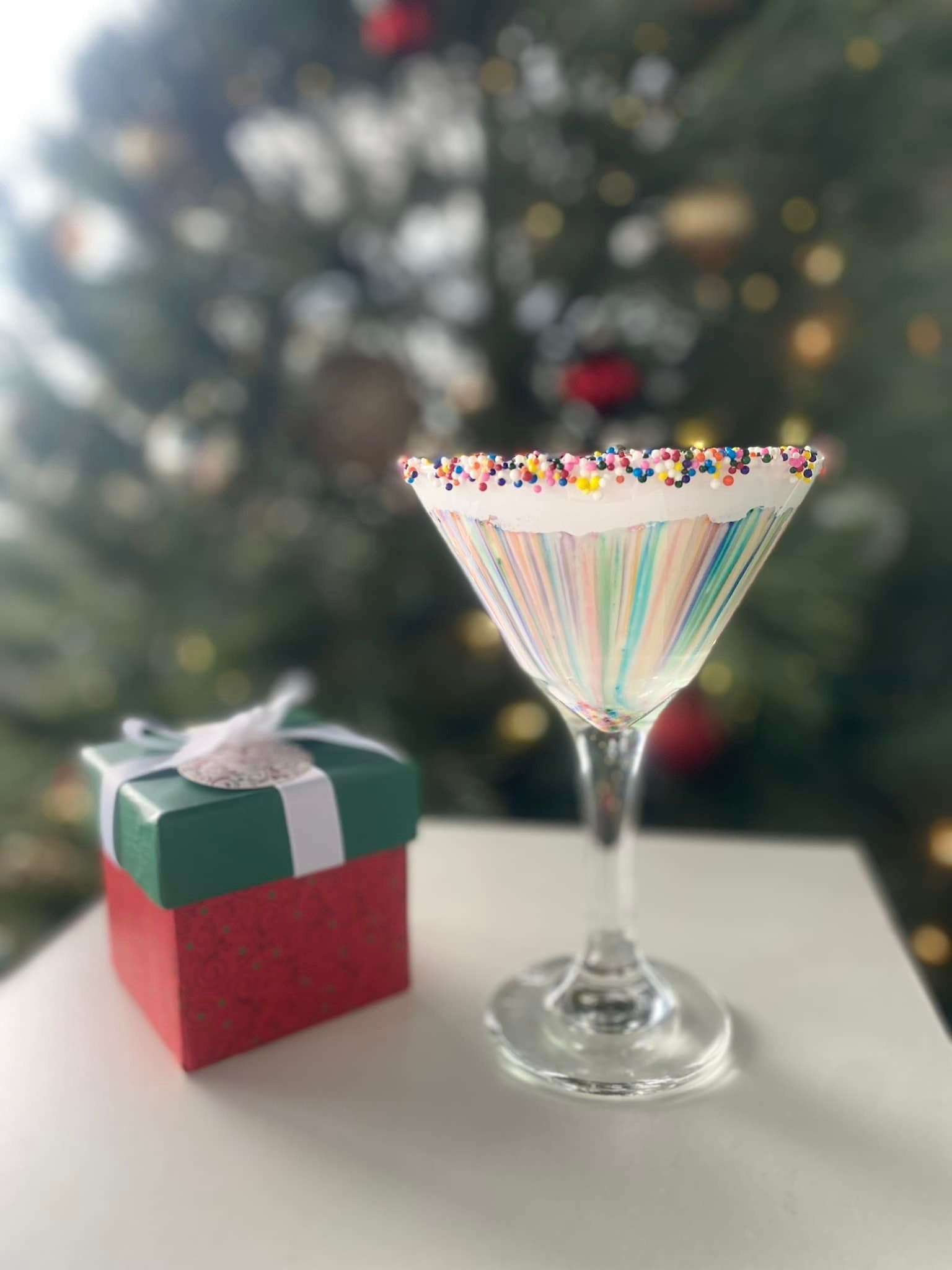 Cocktail glass with sprinkles, a small gift box, Christmas tree in the background.
