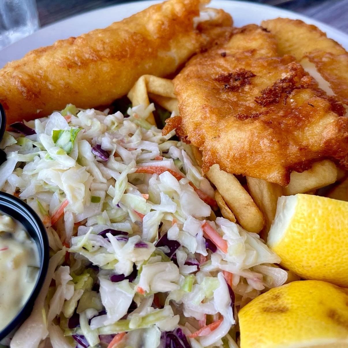 Fish and chips with coleslaw and lemon wedges on a plate, with a side of sauce.
