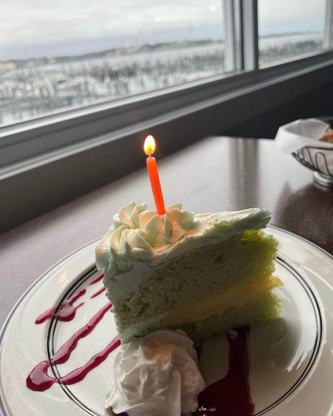 Slice of green cake with a lit candle, white frosting, and berry drizzle, near a window with a snowy view.