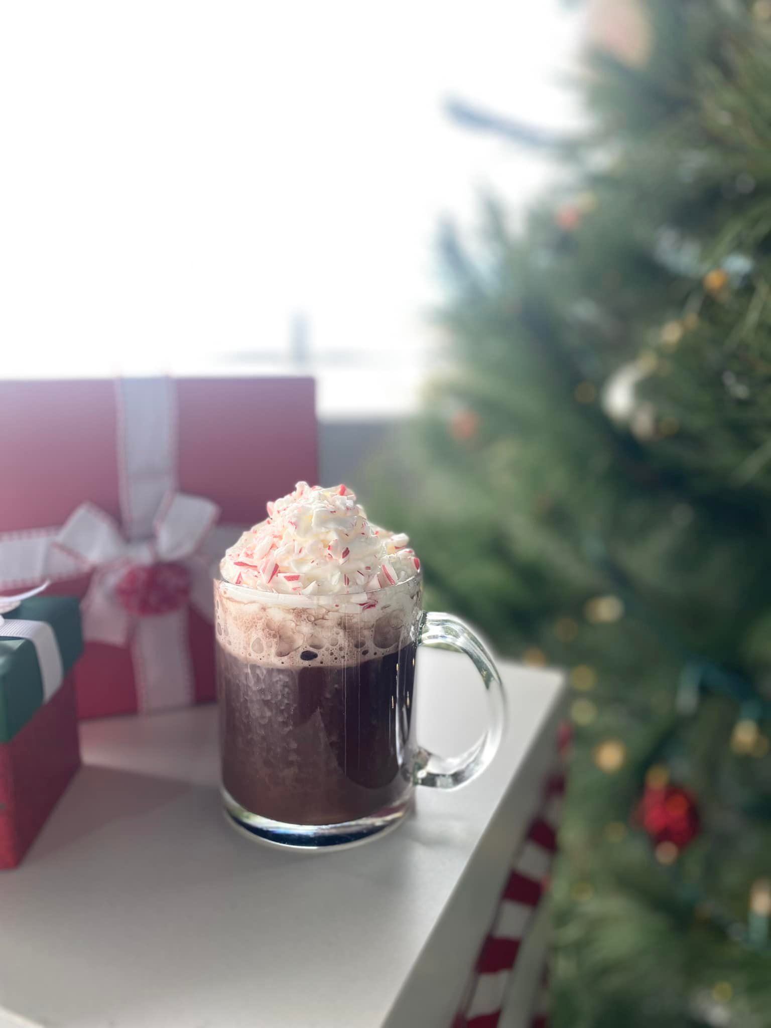 Mug of hot chocolate with whipped cream and crushed candy canes, near presents and a Christmas tree.