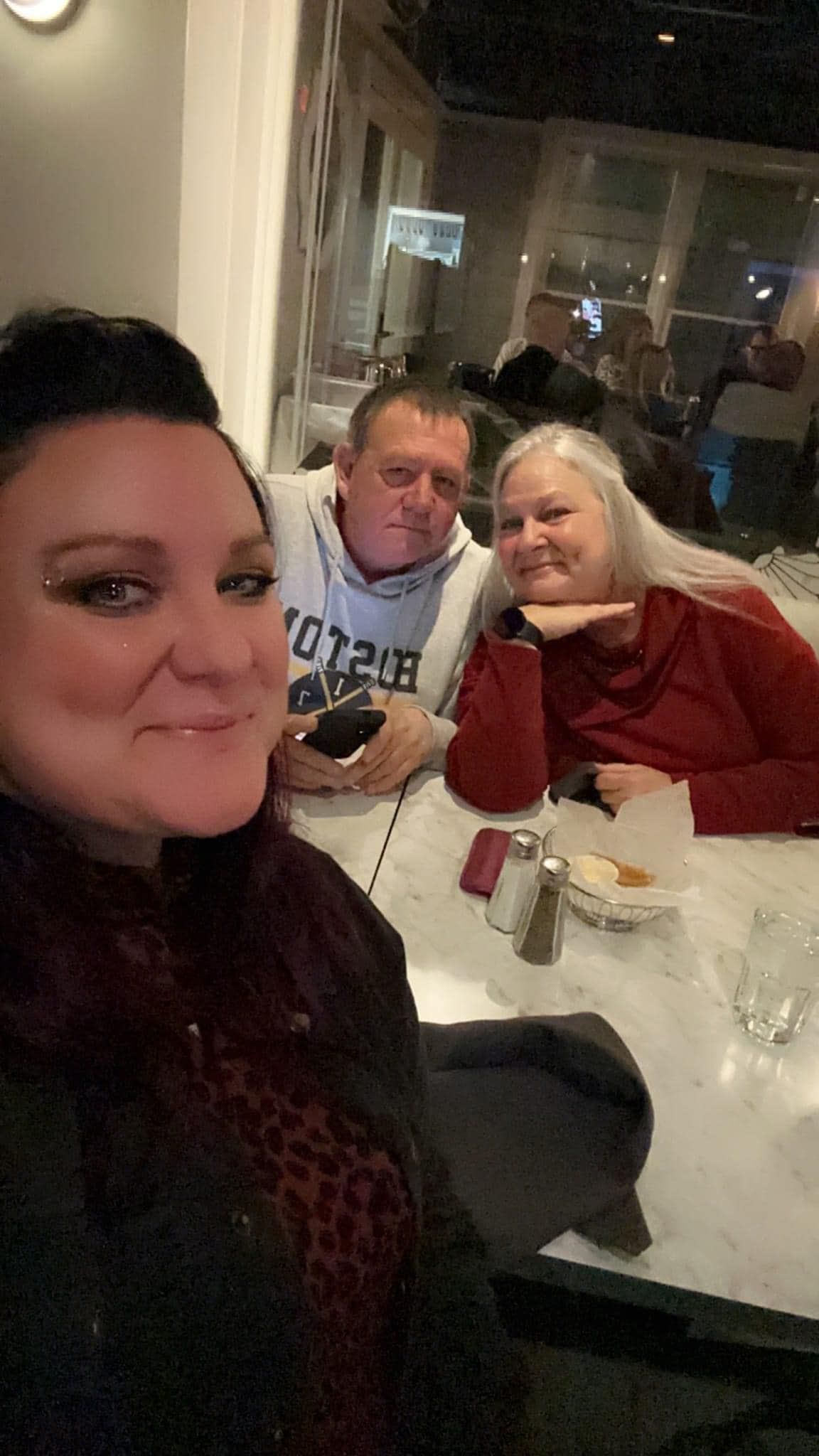 Three people at a restaurant, one taking a selfie. Man in a hoodie, woman in red, other woman smiling with bold makeup.