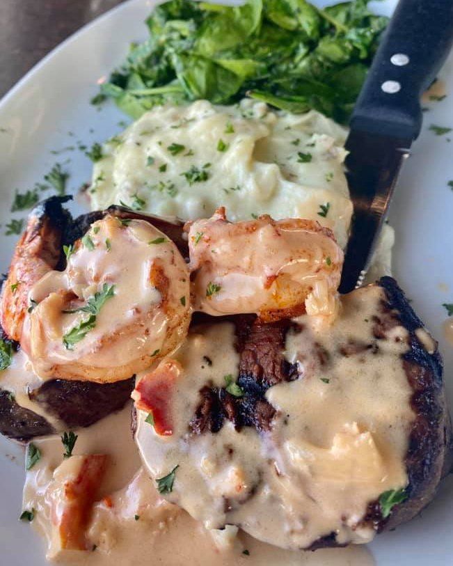 Steak and shrimp dish with creamy sauce, mashed potatoes, and spinach on a white plate.