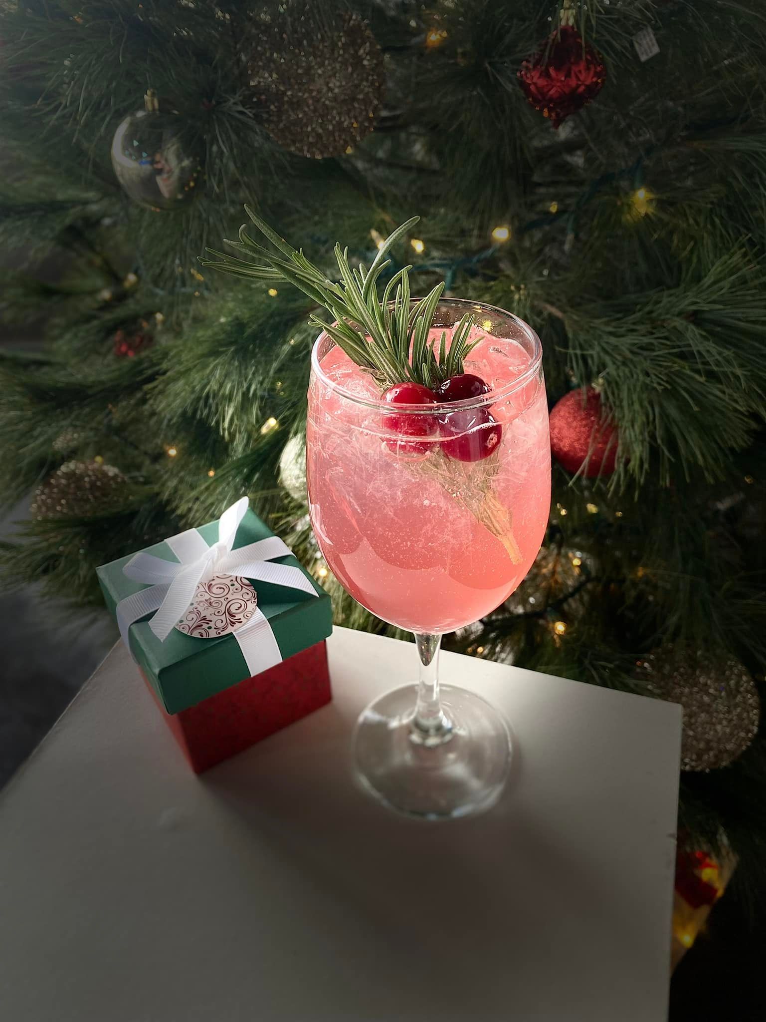 Pink cocktail in a stemmed glass, garnished with rosemary and berries, beside a wrapped gift, in front of a Christmas tree.