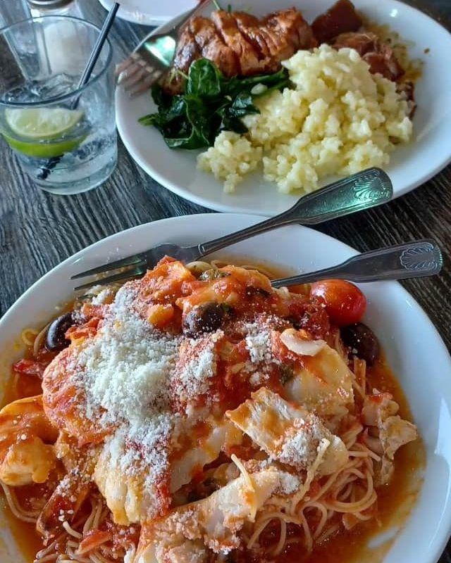 Two plates of food. One with pasta and seafood, the other with meat, vegetables and potatoes.