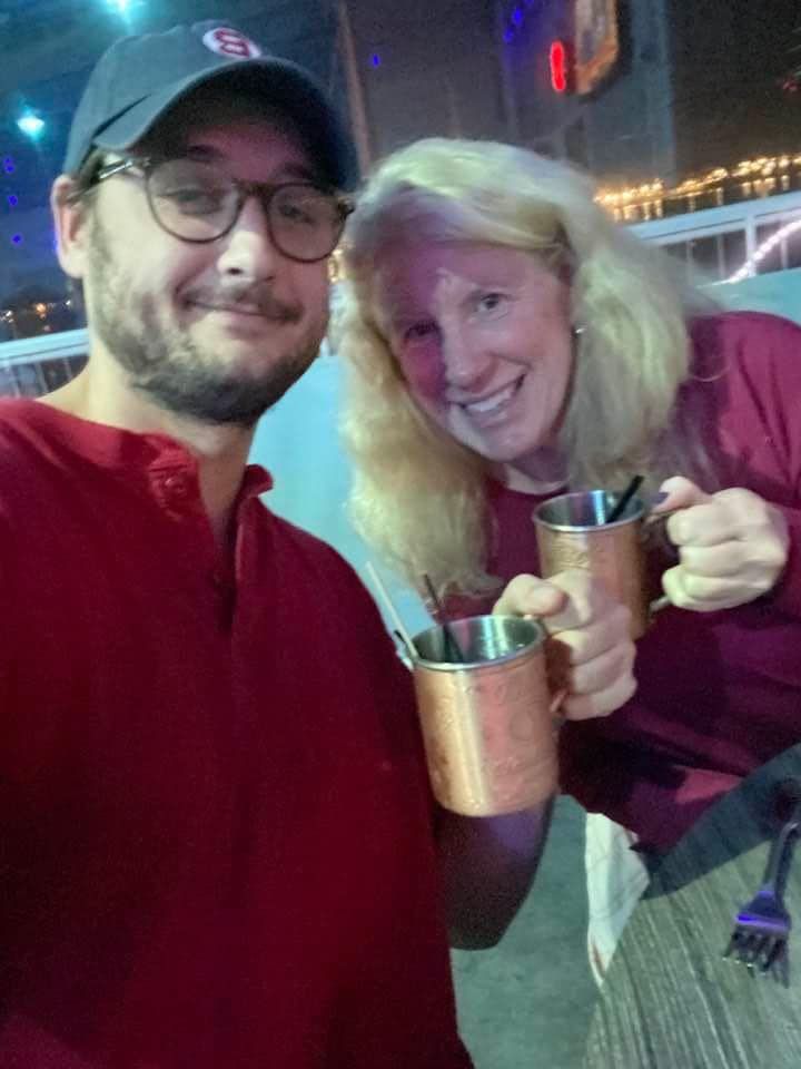 A man and woman smiling, toasting with copper mugs, likely in a bar setting.
