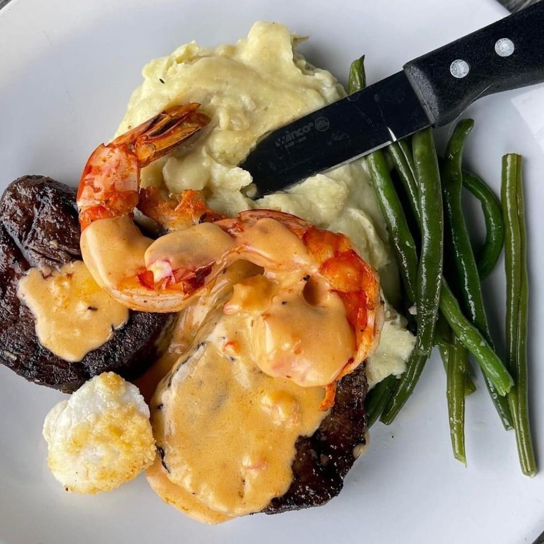 Steak with shrimp and sauce, mashed potatoes, and green beans on a white plate.