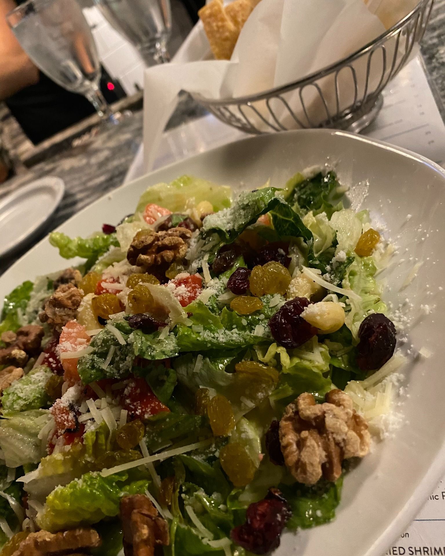 Salad with lettuce, walnuts, raisins, and cheese in a white bowl, bread basket in background.