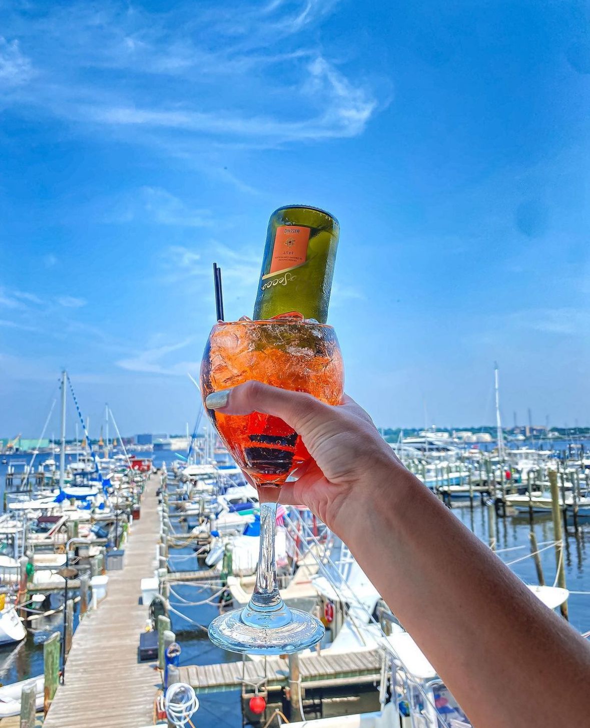 Hand holding a large cocktail with a small bottle, overlooking a marina with boats. Blue sky.