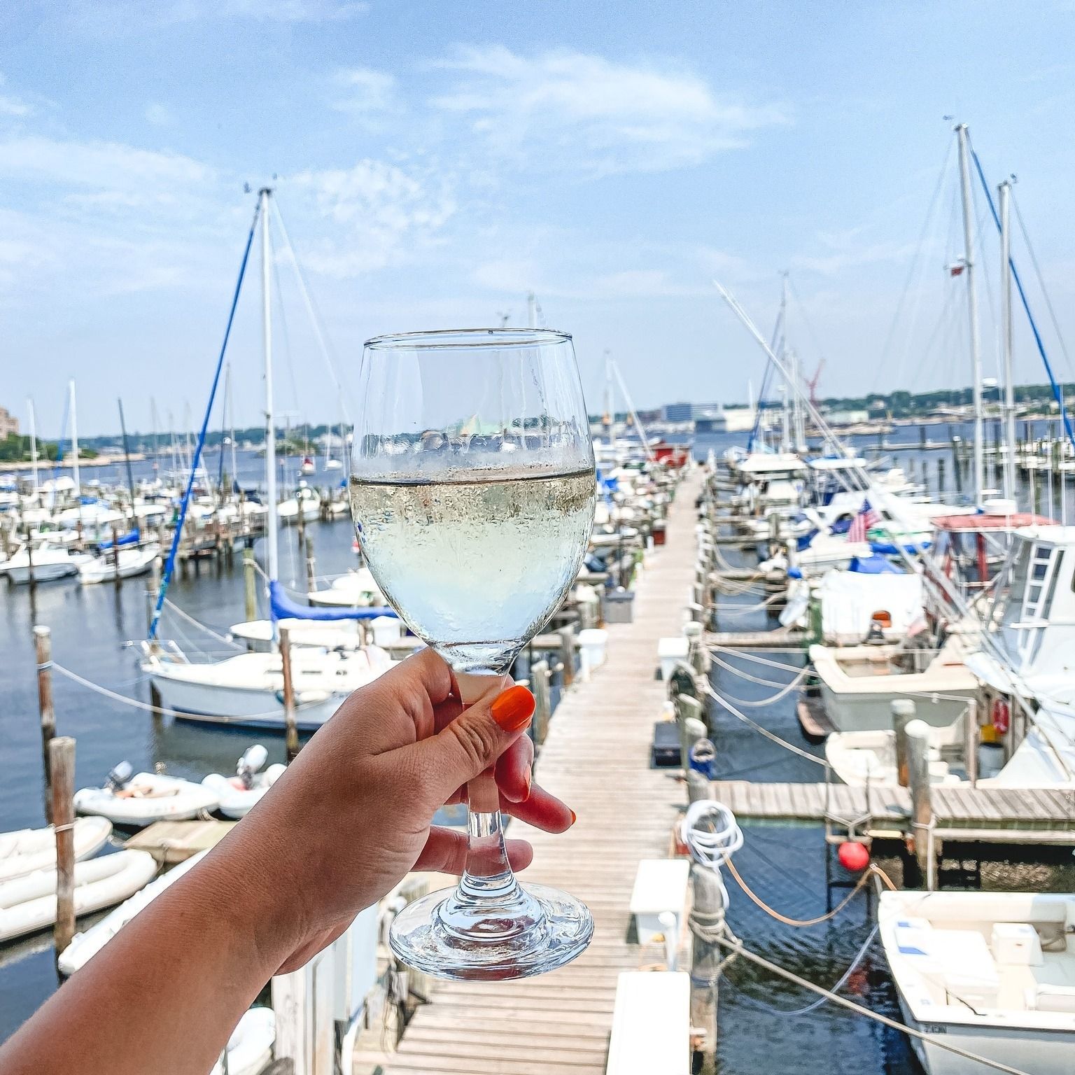 Hand holding a glass of white wine, docks with boats in background. Bright sky and water.
