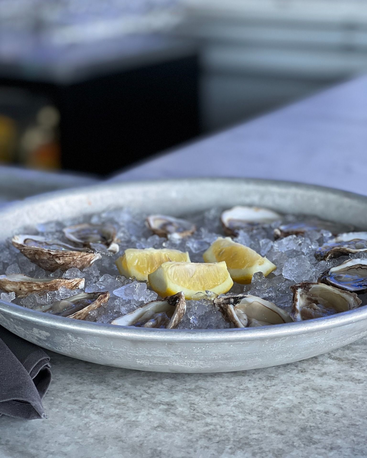 Oysters on ice in a silver tray with lemon wedges, resting on a stone surface.